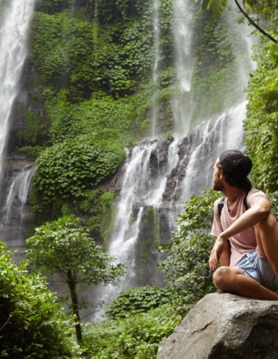 Tourism, travel and adventure. Stylish young hipster sitting on stone with bare feet and turning his head back to see amazing waterfall in background, enjoying happy peaceful moment during holidays